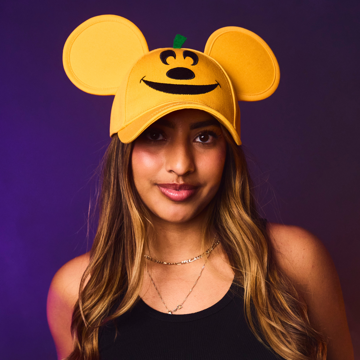 Person wearing an orange cap with a pumpkin design and mouse ears against a purple background