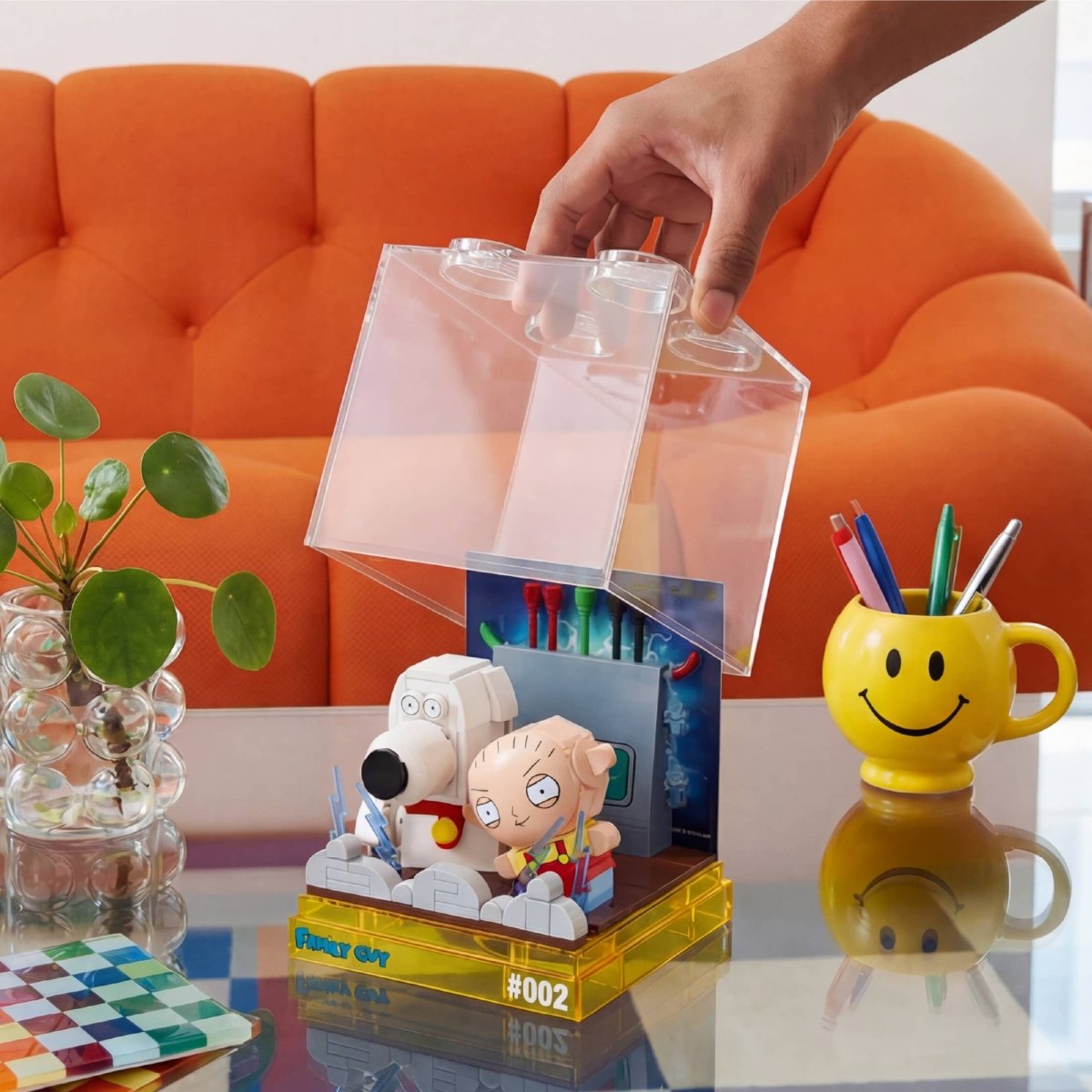 Hand opening a clear plastic box containing small figurines on a table with a colorful mug and plant in the background.