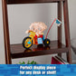 Toy figure of a character on a tricycle on a wooden shelf with a tag, text 'Perfect display piece for any desk or shelf' at the bottom.