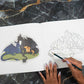 Coloring book with dragon illustration, completed on one side and in progress on the other, on a marble surface.