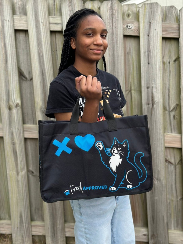 Person holding a black tote bag with a cat design and text outdoors.