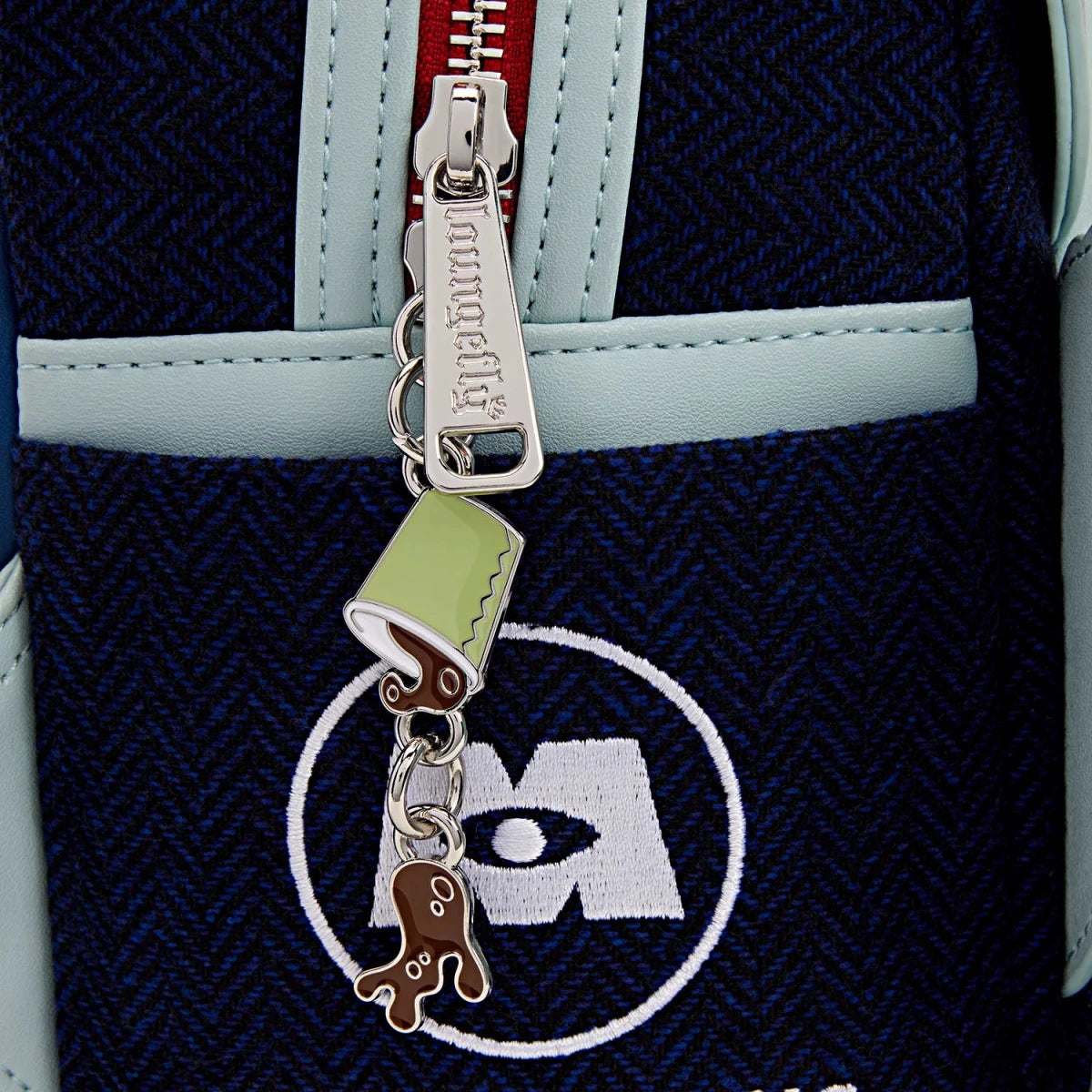Close-up of a dark navy blue herringbone-patterned bag with a silver zipper pull engraved with 'Kikimora', featuring a mint green and brown octopus charm and a white circular logo patch.