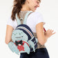 A woman wearing a white t-shirt and navy skirt carries a Disney Pixar Inside Out 2 character backpack on her shoulder. The backpack features a light blue crab character wearing a red polka-dot shirt and black bow tie, with a navy blue top and a checkered strap.