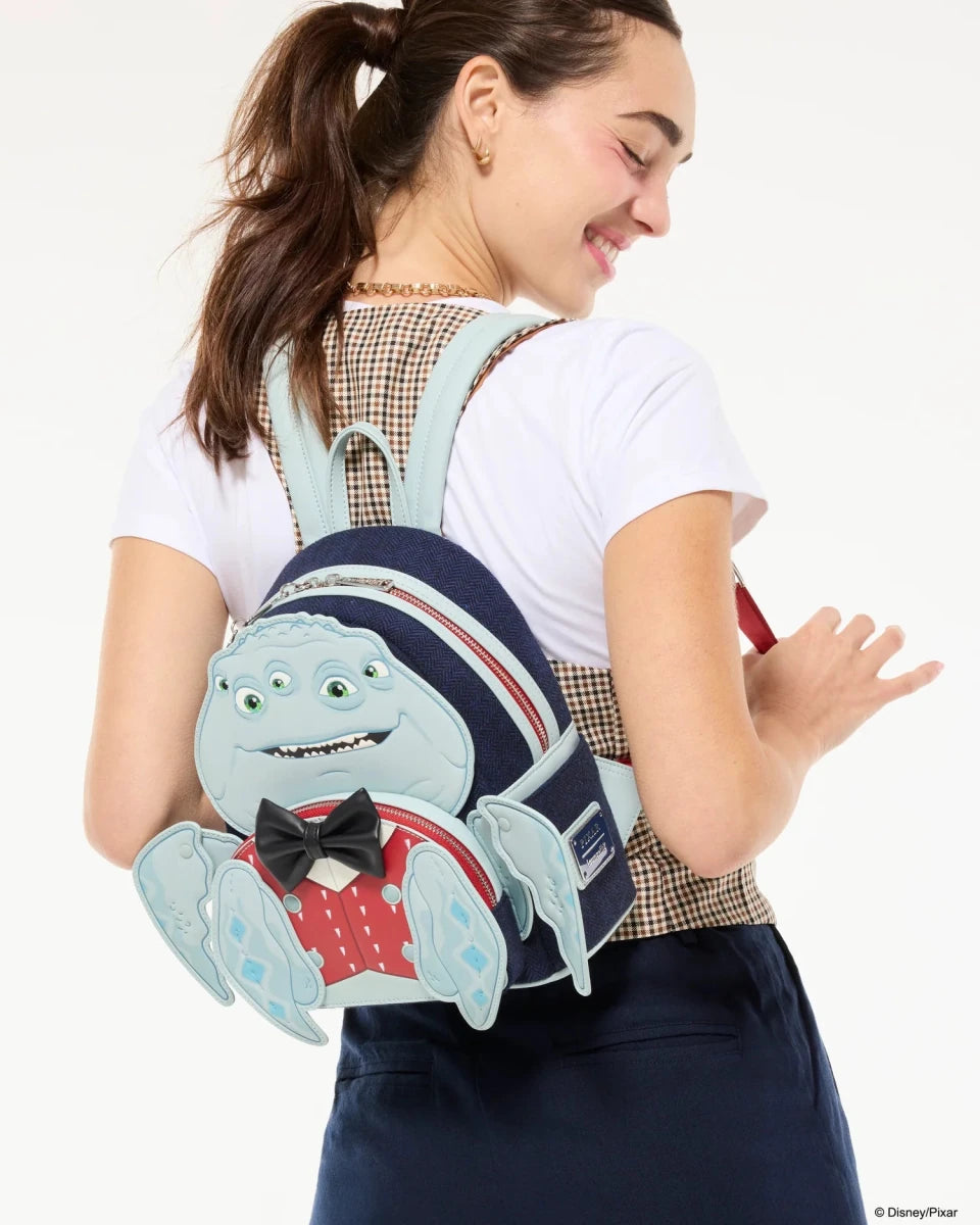 A woman wearing a white t-shirt and navy skirt carries a Disney Pixar Inside Out 2 character backpack on her shoulder. The backpack features a light blue crab character wearing a red polka-dot shirt and black bow tie, with a navy blue top and a checkered strap.