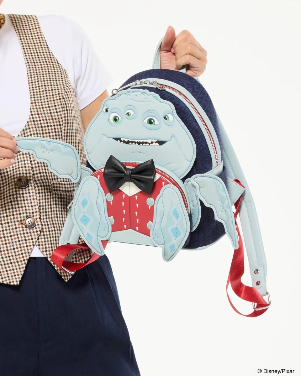 A person holds a small Disney Monsters backpack featuring the character Grommet. The backpack is light blue with a red polka-dot shirt, black bow tie, and dark blue straps, shown against a plain white background.