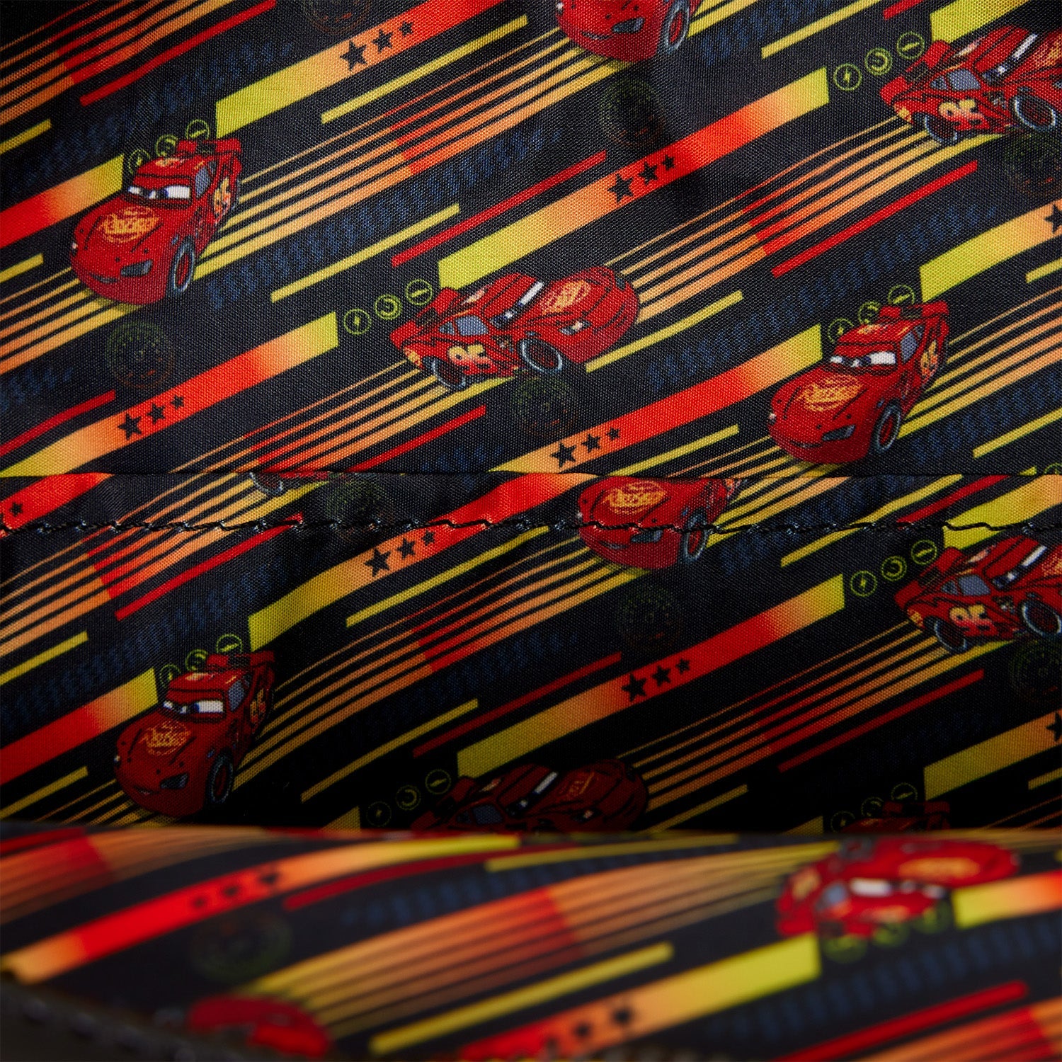 Close-up of a printed fabric featuring repeated red Lightning McQueen car illustrations over a black background with yellow and orange racing stripes and small green circular accents.