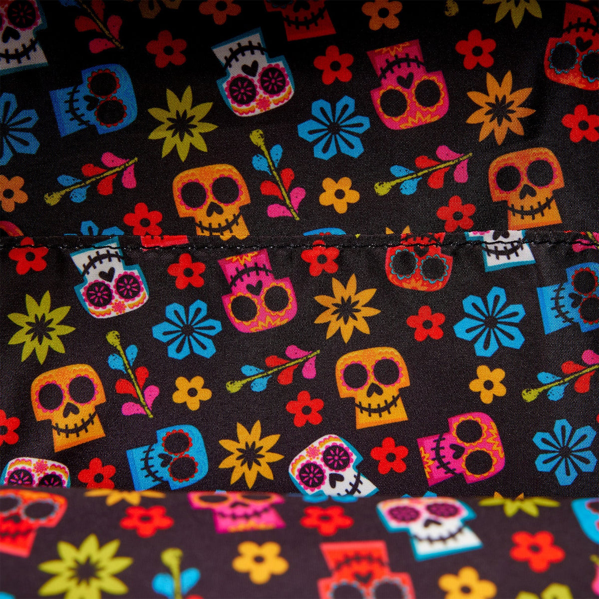 Close-up of a black fabric with a colorful sugar skull and floral pattern, showing a horizontal stitched seam and a folded section of the same fabric at the bottom.