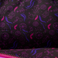 Close-up of a black fabric surface with a repeating pattern of pink and purple whimsical doodles and symbols, including a ghost, swirl, and abstract shapes. A stitched seam runs horizontally across the center, and part of a bright pink zipper is visible along the bottom edge.