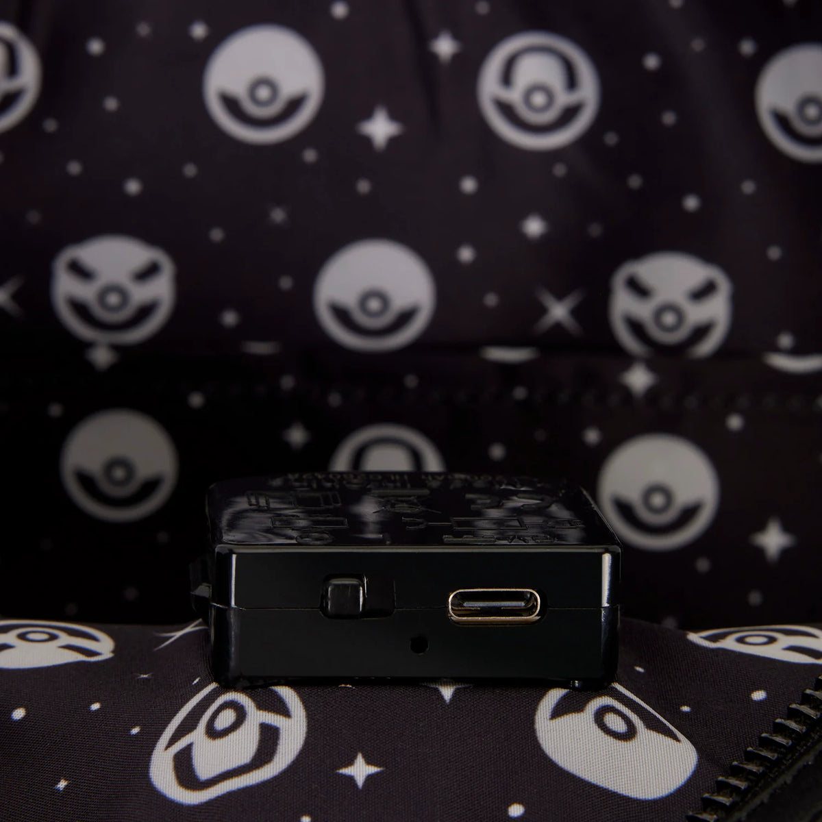A small black rectangular electronic device with a USB-C port, resting on a dark patterned fabric with white Pokéball motifs.