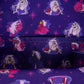 A close-up of a purple fabric printed with cartoon characters in pink, white, and red, including a central character with a green bow and a pink bird-like figure, with a stitched seam visible across the middle.