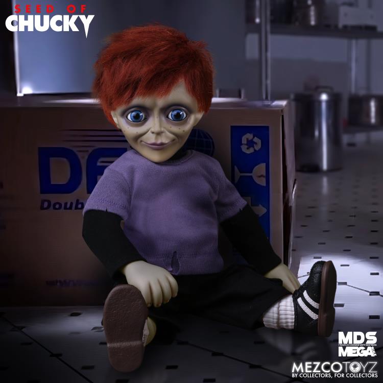 Toy figure of a character with red hair and purple shirt sitting on the floor next to a cardboard box.