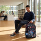 Man sitting in a modern library or study area using a tablet with a colorful backpack next to him.