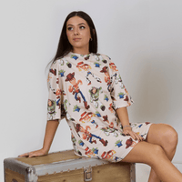 Woman wearing a colorful dress with cartoon character print, sitting on a wooden box against a plain background.