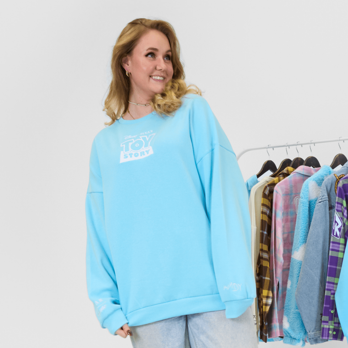 Woman wearing a light blue sweatshirt with a logo, standing next to a rack of clothes.