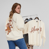 Woman wearing a cream sweatshirt with a cartoon character, standing next to another sweatshirt on a rack.