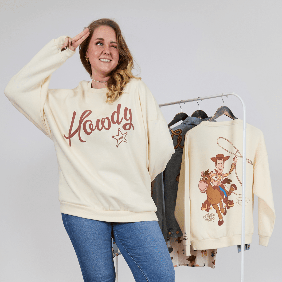 Woman wearing a 'Howdy' sweatshirt with a cowboy graphic, standing next to a rack of similar shirts.