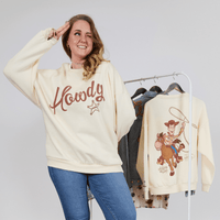 Woman wearing a 'Howdy' sweatshirt with a cowboy graphic, standing next to a rack of similar shirts.