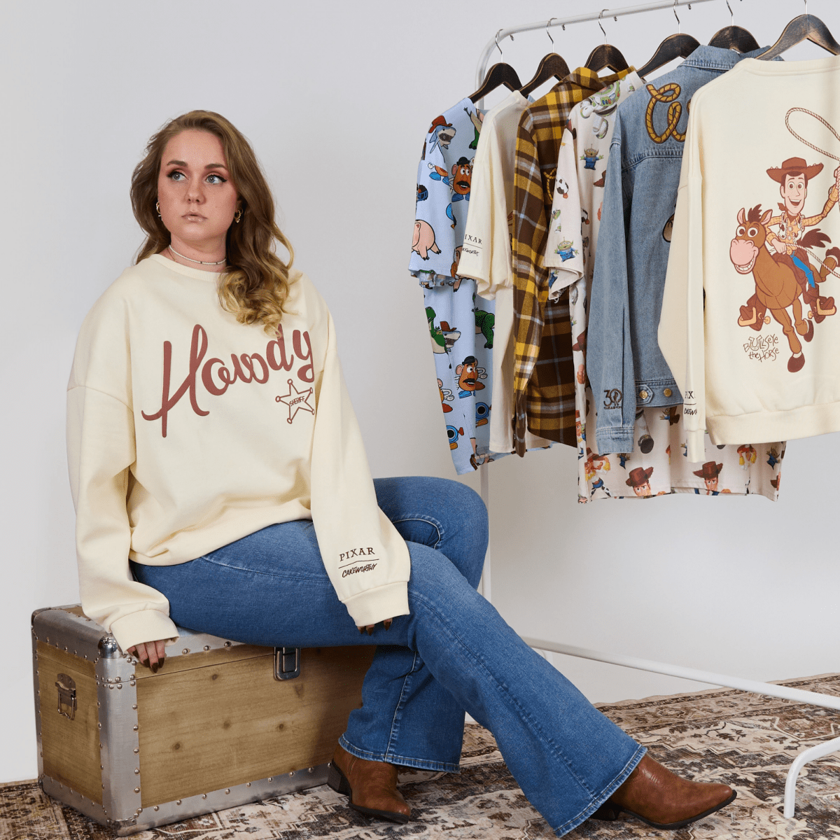 Woman wearing a 'Howdy' sweatshirt sitting on a suitcase with a rack of clothes in the background