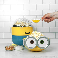 Minion-themed popcorn maker with a hand pouring oil into it, set against a white tiled wall.