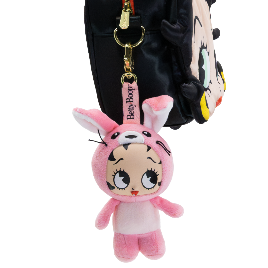 A pink Betty Boop plush bunny keychain charm hanging from a black bag with a cartoon face. The plush features a pink bunny hood with long ears, large expressive eyes, and a pink belly, with a pink tag reading 'Betty Boop' and gold-tone hardware.