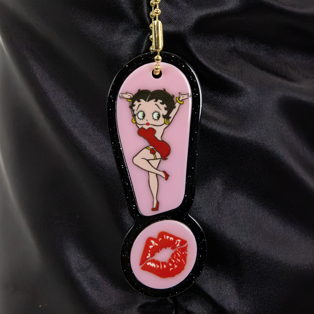 A Betty Boop themed keychain featuring a pink exclamation mark pendant with a cartoon Betty Boop figure and a round lip emblem, hanging against a black satin fabric background.