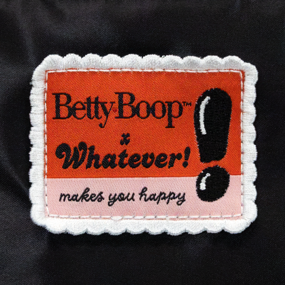 An embroidered patch with a scalloped white border, featuring the text 'BettyBoop x Whatever! makes you happy' in black on orange and light pink sections, set against a black fabric background.