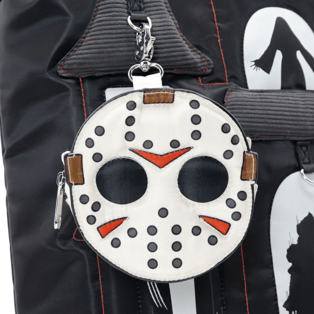 A round white faux-leather bag charm designed like a hockey mask, featuring black polka dots, black eye holes, red triangular mouth details, and brown leather accents, hanging from a silver clasp on a black backpack.