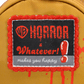 A close-up of a yellow fabric item, likely a backpack, featuring a decorative embroidered patch. The patch has a red frayed border, a black center, and red and white embroidery that reads 'HORROR x Whatever! makes you happy' alongside the red WB shield logo.