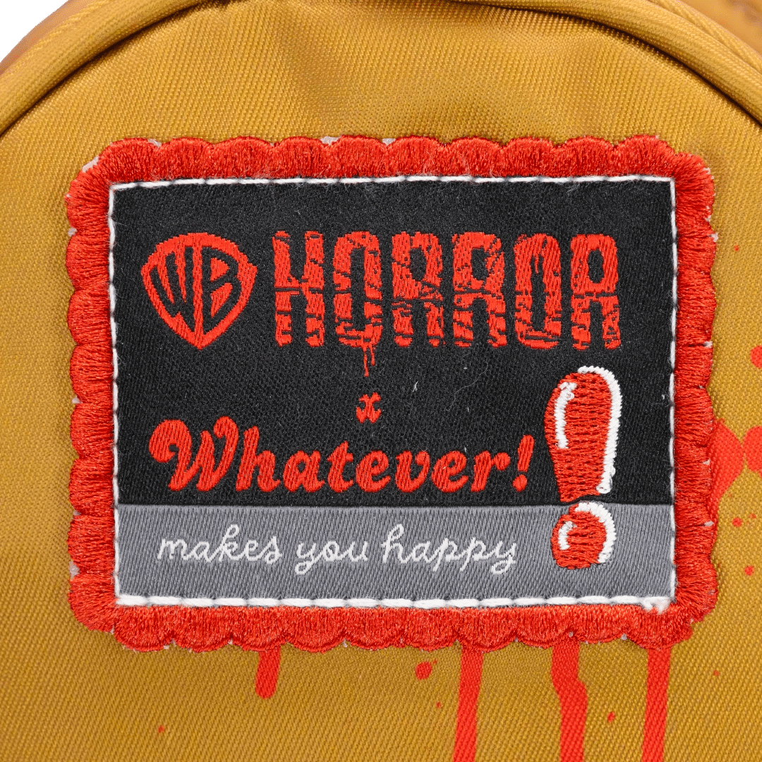 A close-up of a yellow fabric item, likely a backpack, featuring a decorative embroidered patch. The patch has a red frayed border, a black center, and red and white embroidery that reads 'HORROR x Whatever! makes you happy' alongside the red WB shield logo.