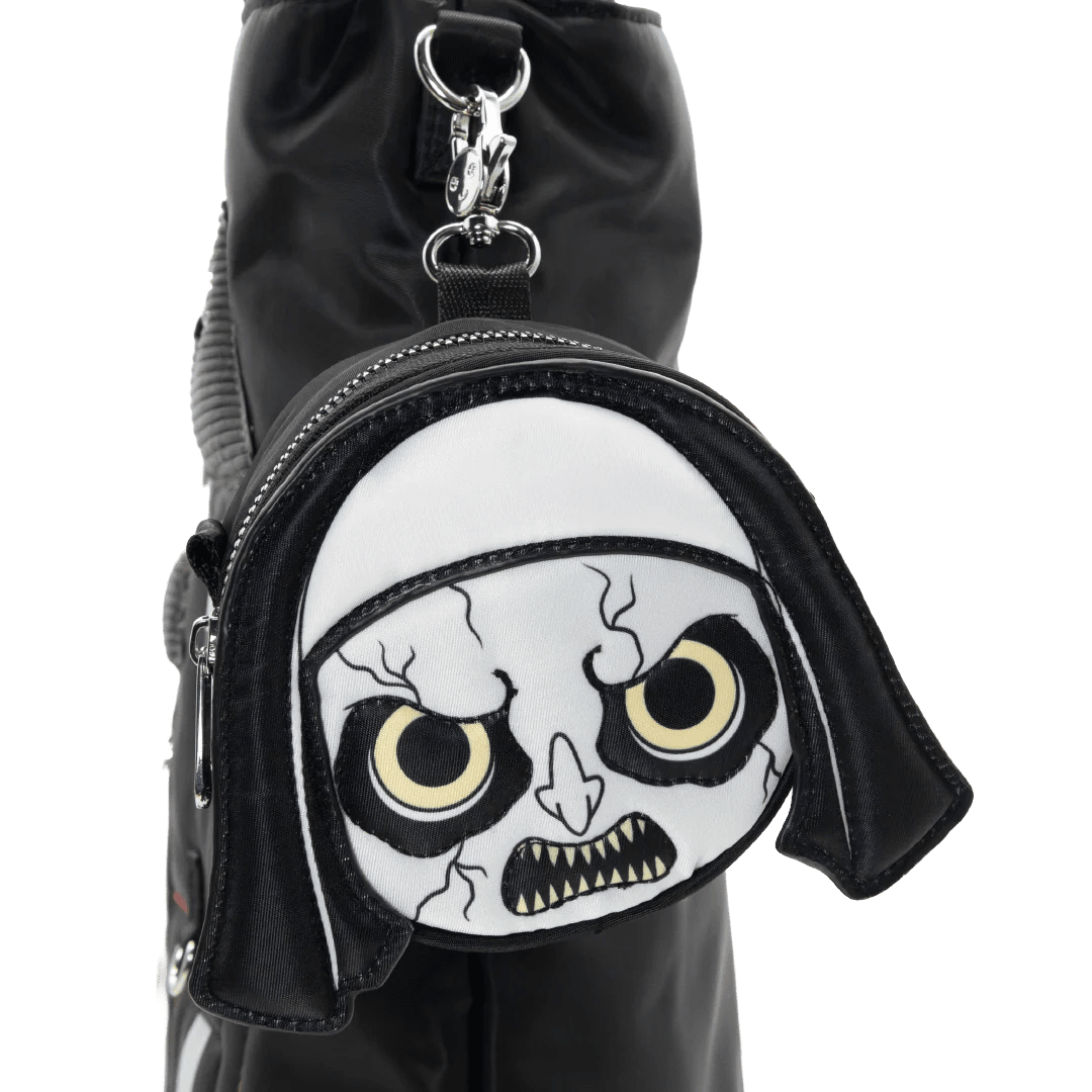 A black nylon shoulder bag featuring a round front zipper pocket with a white fabric patch shaped like a cartoon skull face with yellow eyes and black stitched details, attached to a silver metal clasp.