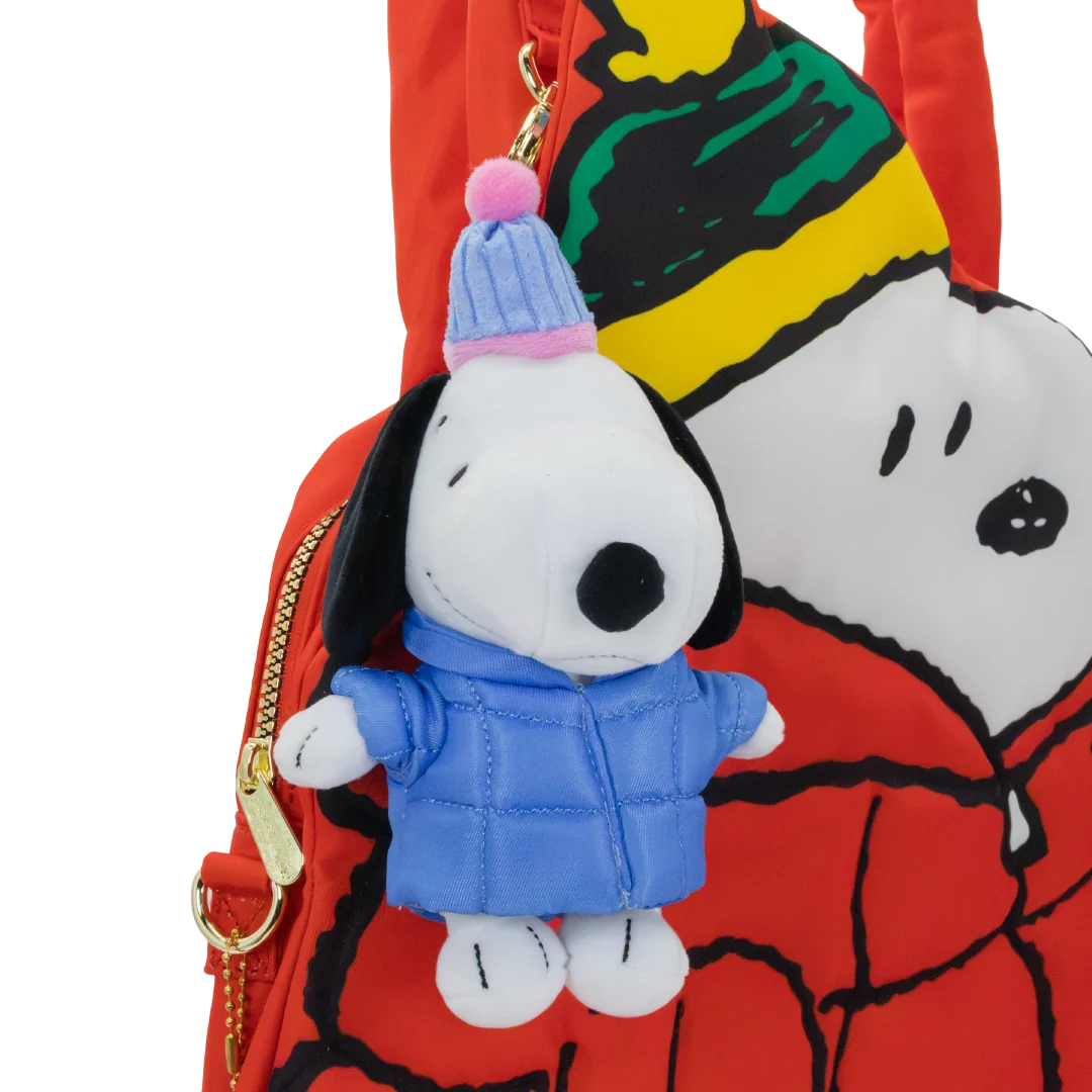 A red Snoopy backpack featuring a plush Snoopy keychain in a blue winter jacket and striped beanie, with a large printed Snoopy face on the back.