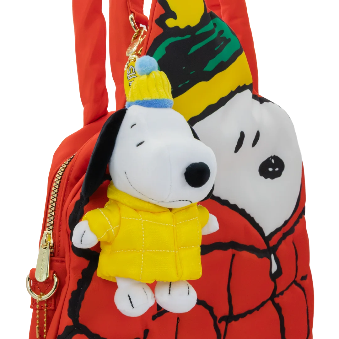 A red Snoopy-themed shoulder bag featuring a plush Snoopy character charm wearing a yellow jacket and blue-and-yellow beanie, shown against a plain white background.