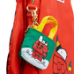A Snoopy-themed mini tote bag keychain charm hanging from a red backpack. The charm features a green body with yellow handles, a white snowy base, and an illustration of Snoopy on a red doghouse wrapped in Christmas lights.