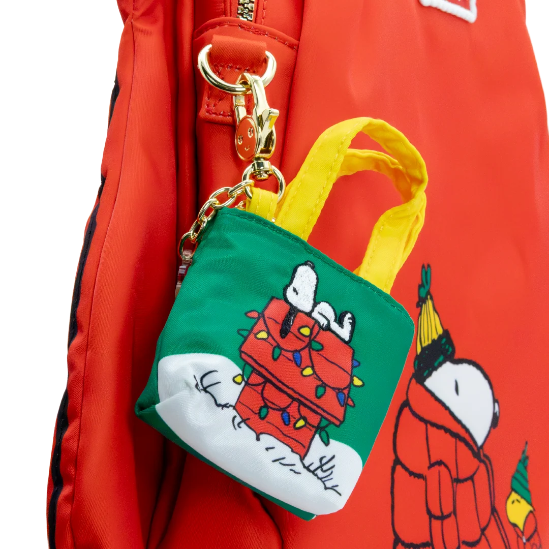 A Snoopy-themed mini tote bag keychain charm hanging from a red backpack. The charm features a green body with yellow handles, a white snowy base, and an illustration of Snoopy on a red doghouse wrapped in Christmas lights.