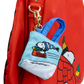 A red nylon backpack with a Snoopy winter-themed mini tote bag charm attached by a gold clasp. The charm features a light blue top with embroidered Snoopy sledding on snow, and the red backpack has a cartoon character print on the side.