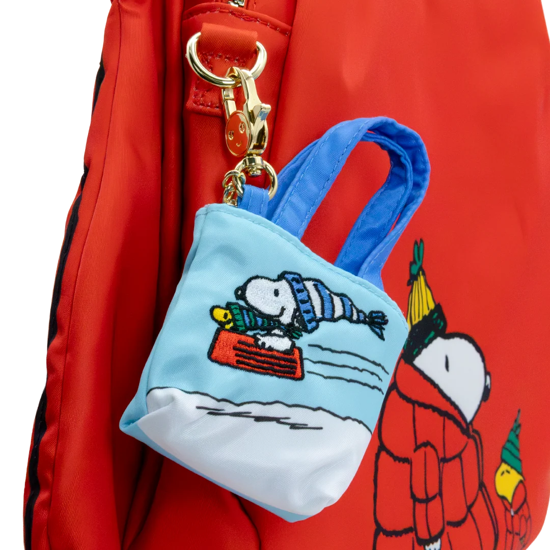 A red nylon backpack with a Snoopy winter-themed mini tote bag charm attached by a gold clasp. The charm features a light blue top with embroidered Snoopy sledding on snow, and the red backpack has a cartoon character print on the side.