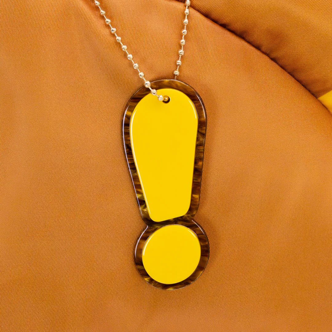 Neon yellow exclamation mark pendant on a chain against an orange fabric background