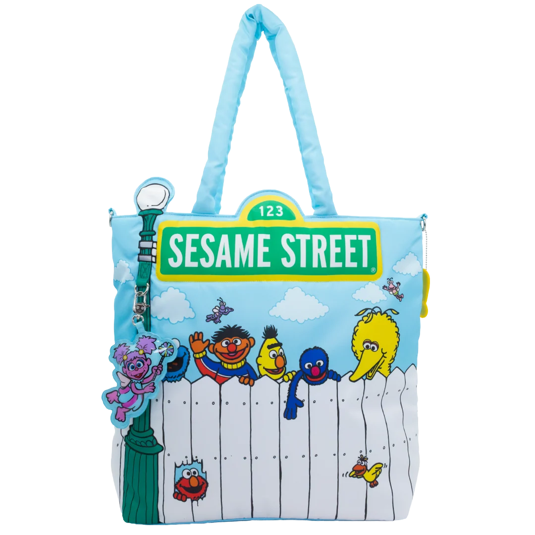 A light blue and white Sesame Street tote bag featuring characters including Big Bird, Oscar the Grouch, Tweety, and Elmo under a green 'Sesame Street' street sign, with a green street lamp charm attached.