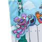 A close-up of a colorful zippered pouch featuring a cartoon character charm. The pouch has a light blue sky and white fence print, with a silver zipper and a clear acrylic charm of a pink and purple fairy holding a wand.