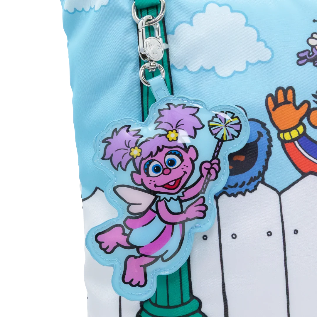 A close-up of a colorful zippered pouch featuring a cartoon character charm. The pouch has a light blue sky and white fence print, with a silver zipper and a clear acrylic charm of a pink and purple fairy holding a wand.