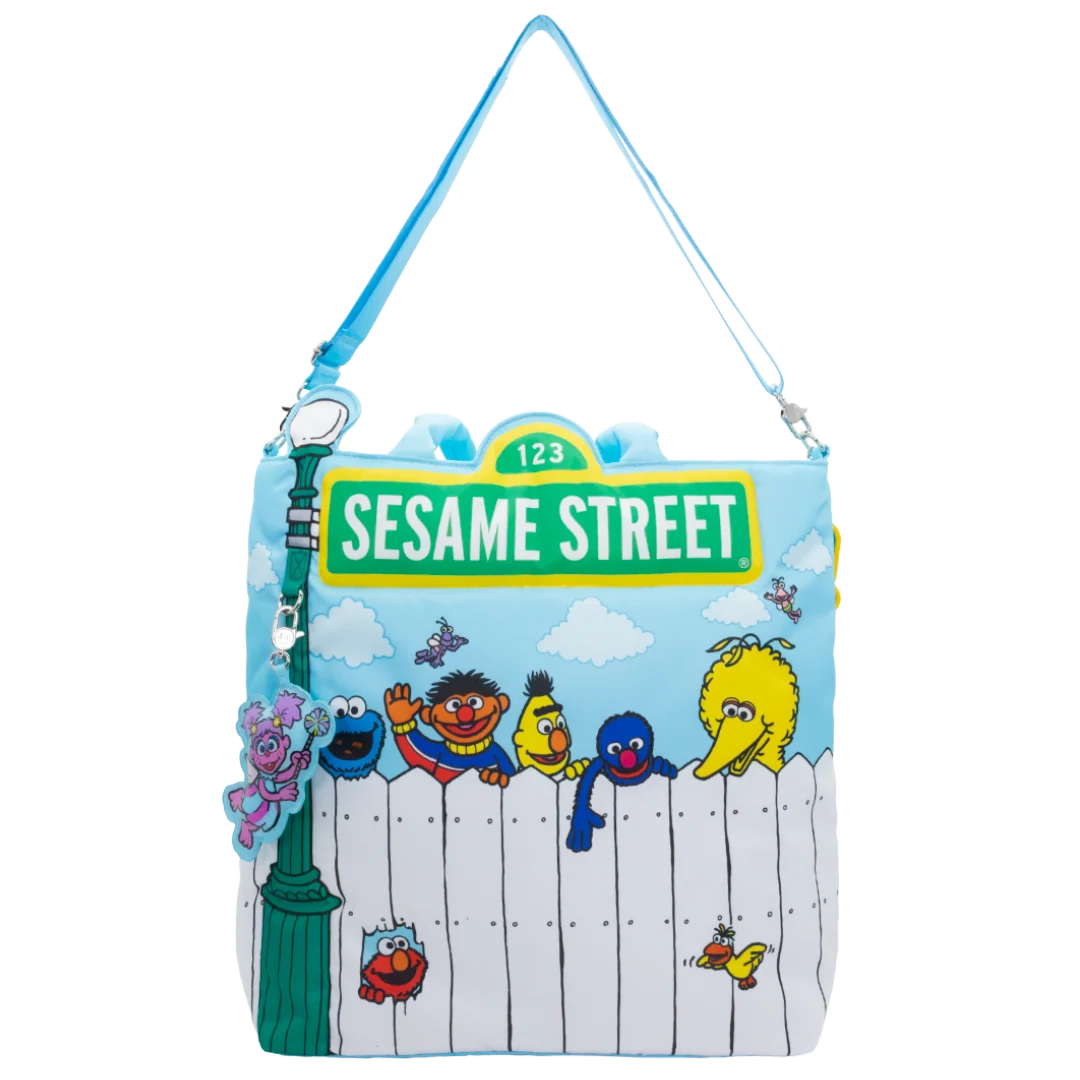 A Sesame Street themed shoulder bag featuring characters like Elmo, Cookie Monster, Big Bird, and Bertie behind a white picket fence on a light blue sky background, with a green '123 SESAME STREET' sign and a blue strap.