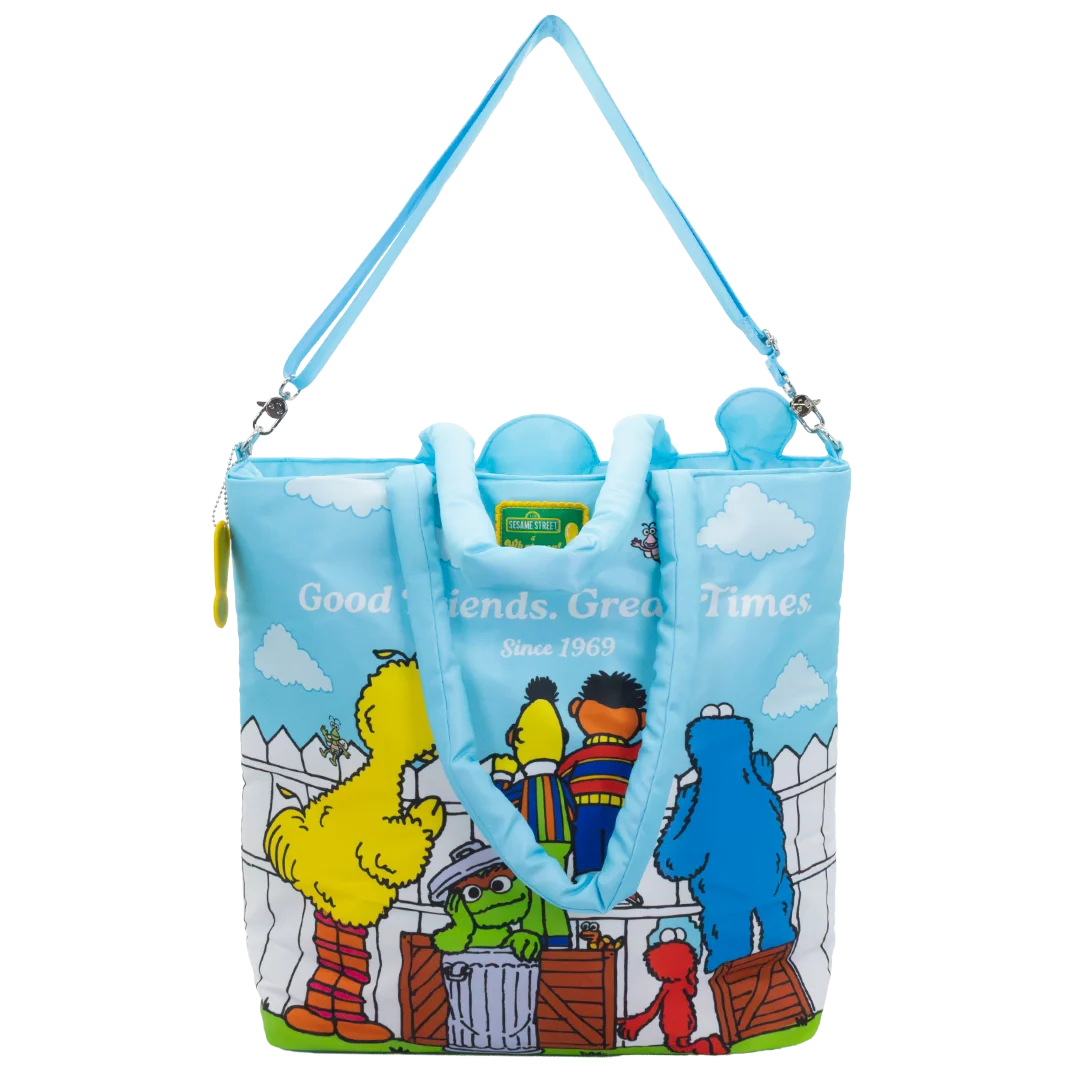 A light blue tote bag featuring a colorful print of Sesame Street characters, including Big Bird, Cookie Monster, and the Blue Monster, with the text 'Good Friends, Great Times Since 1969'. The bag has a light blue shoulder strap and handles, and a Sesame Street logo is visible on the front.