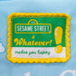 A close-up of a Sesame Street embroidered patch on a light blue fabric background. The patch features the classic green and yellow Sesame Street logo with the phrase 'Whatever! makes you happy' in green and yellow embroidery, surrounded by a yellow scalloped border.