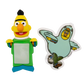Two small character-themed items on a plain white background: a yellow puppet-like figure with a green torso and blue feet holding a blank white panel, and a die-cut sticker or decal of a light blue bird character with a yellow face.