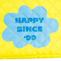 Yellow quilted pillow with blue flower-shaped patch displaying 'Happy Since '99'.