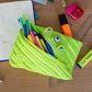 Green monster-shaped pencil case with stationery items on a wooden desk