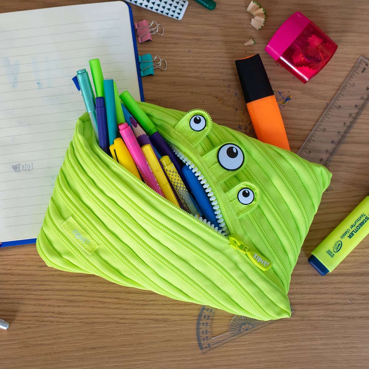 Green monster-shaped pencil case with stationery items on a wooden desk