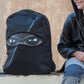 Person sitting next to a black backpack with a hood and face design on a stone wall.