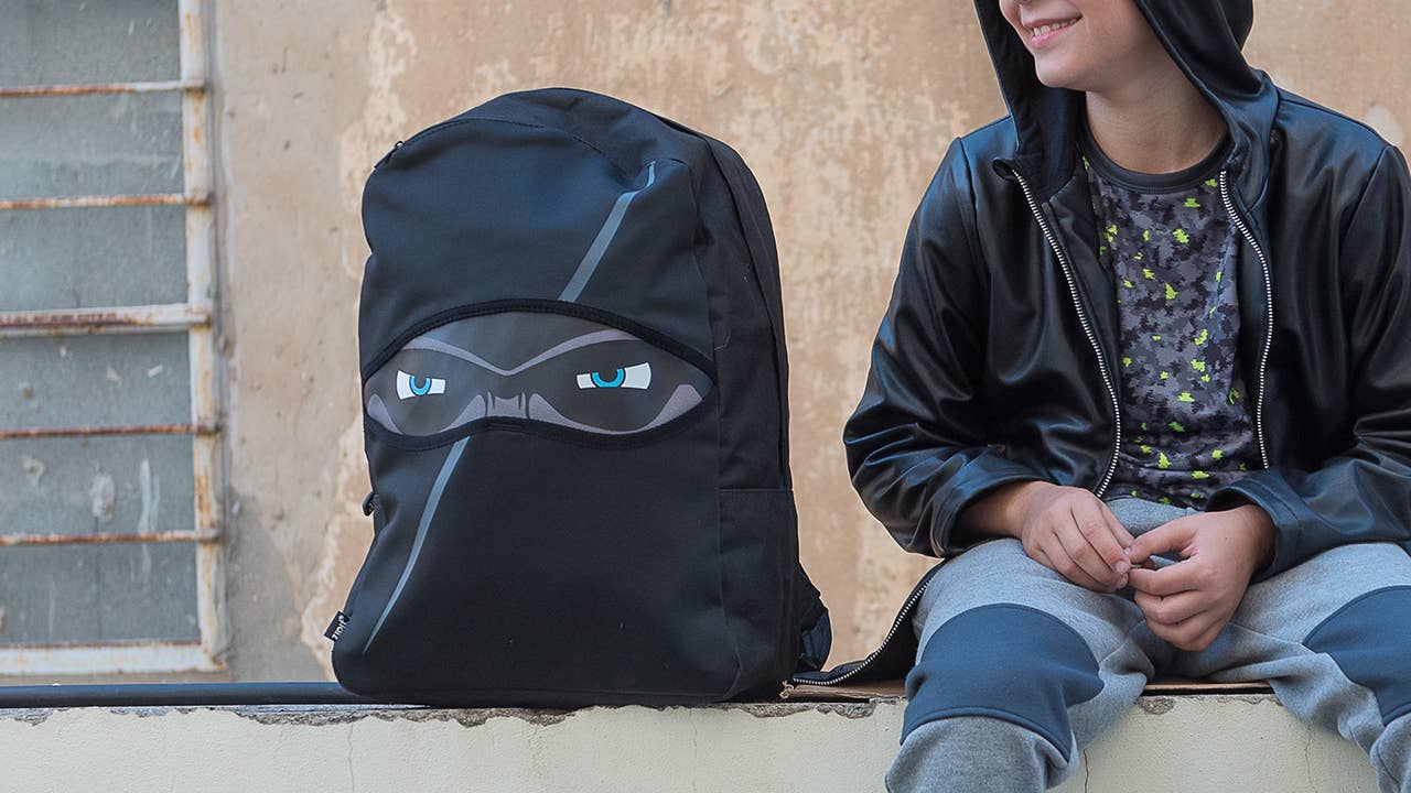 Person sitting next to a black backpack with a hood and face design on a stone wall.