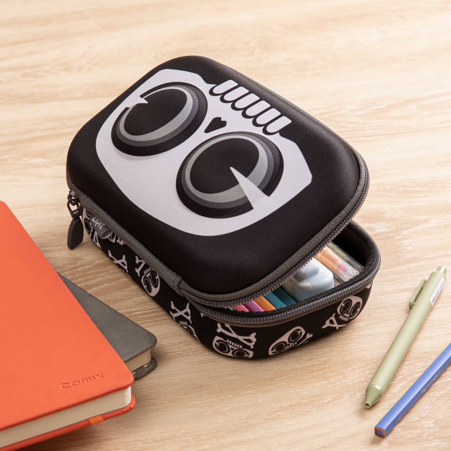 Black pencil case with white logo on a wooden surface with stationery items.